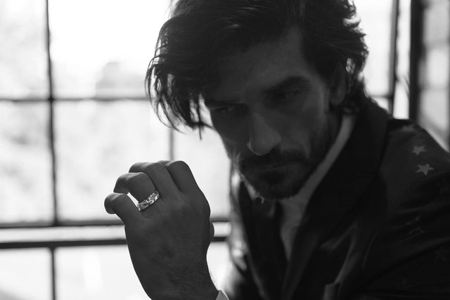A black and white photo of a man wearing a black jacket in front of a large window. He is wearing a platinum stephen einhorn band